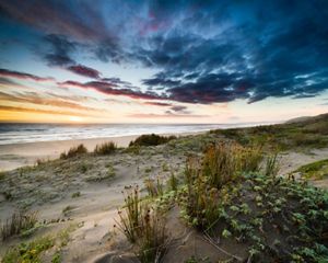 Dramatic landscape view of sand dunes overlooking a beach and ocean under a cloud-filled sky colored in blue and orange.